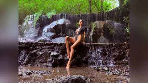 Woman in a black swimsuit sitting on rocks by a lush tropical waterfall in a serene jungle setting.