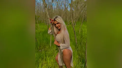 Anita stands in a grassy field, wearing a white mesh bodysuit while leaning against a thin tree trunk.