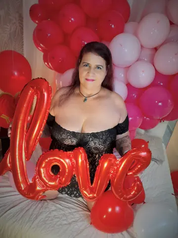 LaurenRiley sits on a bed wearing a black lace bodysuit, posing behind a large red LOVE balloon surrounded by red and pink balloons.