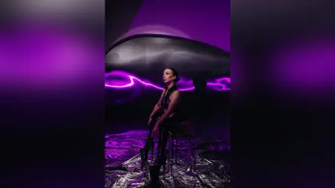 Woman in edgy black outfit sitting in a futuristic room with neon purple lighting and metallic floor decor.