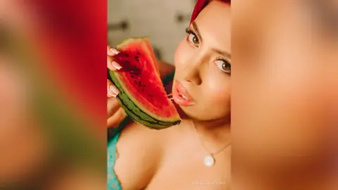 IsabellaEstrada holds a slice of red watermelon near her lips, showing her face, hair, and cleavage in a blue top.