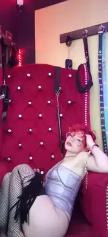 Nyxaria poses in a shiny lavender bodysuit and fishnet stockings while lounging on a red tufted chair.