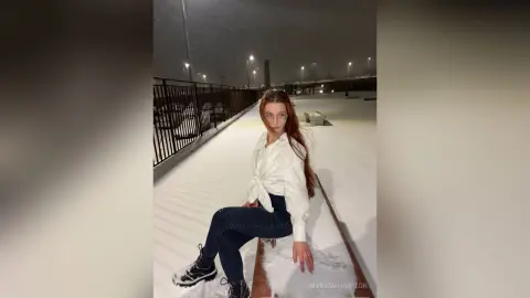 Young woman in a white blouse and dark jeans sitting on a bench in a snowy outdoor setting at night.
