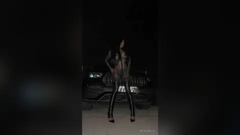 NicoleBella stands naked in a black sheer bodysuit and thigh-high boots while posing in front of a black car at night.