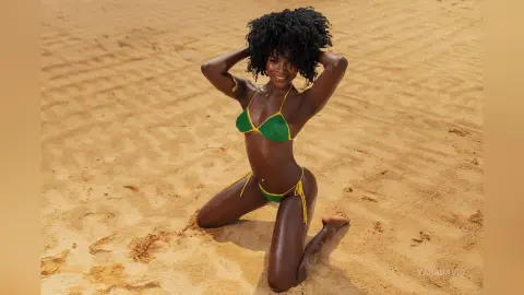 Smiling Black woman in a green crochet bikini posing on a sunny sandy beach.