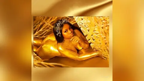 KeianHoz poses naked with gold paint covering her body and a white beaded headpiece.