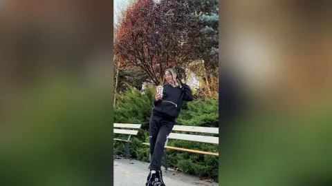 Young woman in black casual outfit holding a drink while sitting on a park bench near autumn trees.