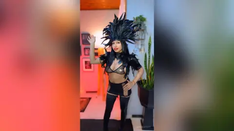 Woman in black feathered cosplay costume and thigh-high boots posing in a modern living room.