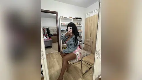 Young woman in a denim jacket and floral skirt taking a mirror selfie in a bright bedroom setting.
