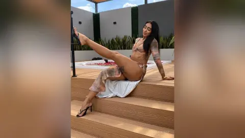 Stylish woman in a beige bikini and white skirt relaxing on wooden stairs at a sunny outdoor rooftop pool.