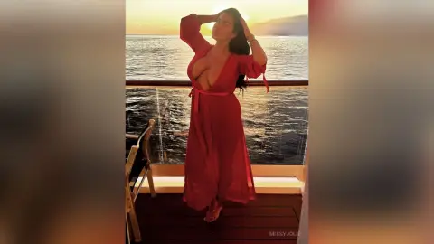 MissyJolie stands on a balcony overlooking the ocean wearing a long red dress with a deep plunging neckline.
