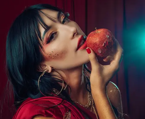 Glamorous woman with dark hair and red makeup biting a glittery red apple in a moody, dramatic setting.