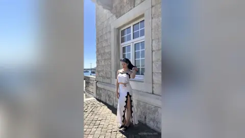 Elegant woman in a white sleeveless maxi dress standing by a stone building on a sunny day.