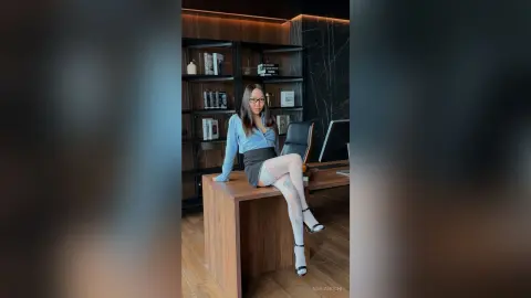 Stylish woman in a light blue blouse and black skirt sitting on a desk in a modern professional office setting.