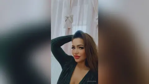 Glamorous woman in a dark green ribbed top posing against white curtains in a bright, airy room.