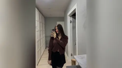 Young woman in a brown sweater taking a mirror selfie in a minimalist modern hallway.