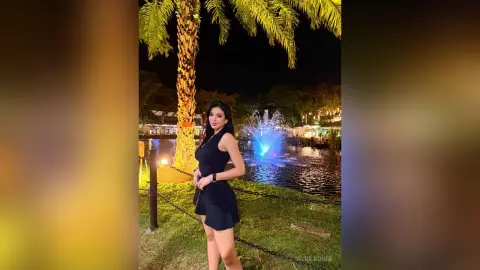 Elegant woman in a black sleeveless dress standing by a glowing fountain at a night garden resort.