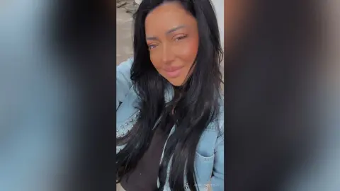 Smiling woman with long dark hair wearing a light blue denim jacket in a bright outdoor setting.