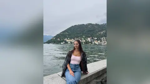 Young woman in a plaid shirt and blue jeans sitting by a scenic lake and mountains under a cloudy sky.