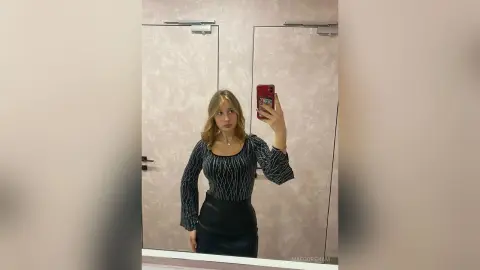 Young woman in a black long-sleeve patterned top and leather skirt taking a mirror selfie in a modern room.