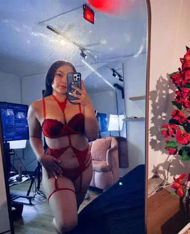Curvy woman in red lace lingerie posing for a mirror selfie in a cozy, dimly lit bedroom.