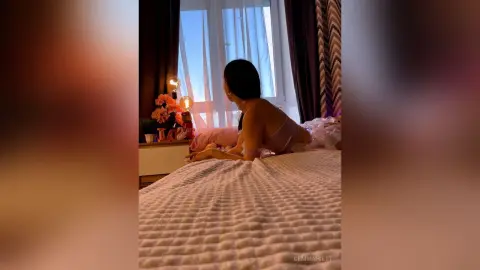 Young woman in a pink silk slip dress relaxing on a bed in a cozy, sunlit bedroom.