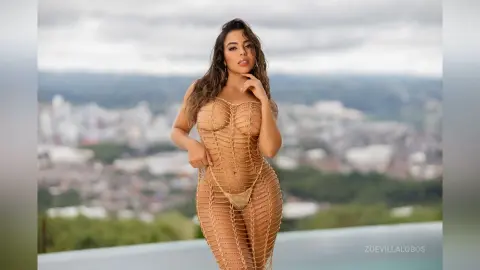 Woman in a sheer gold mesh bodysuit standing on a rooftop overlooking a scenic city landscape at dusk.