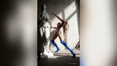 LeslyAnderson poses in a blue strap bikini and matching thigh-high boots next to a white statue in a bright room.