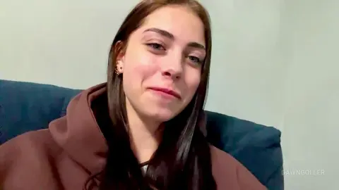 Smiling young woman with long dark hair wearing a brown hoodie sitting on a blue sofa in a bright room.