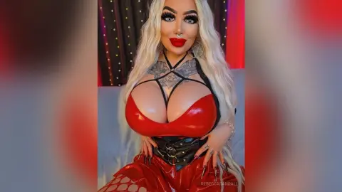 Glamorous blonde woman in a red latex outfit and black harness posing with bold makeup.