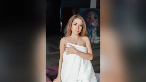 Blonde woman in a white wrap sitting in a moody, dimly lit room with artistic decor.