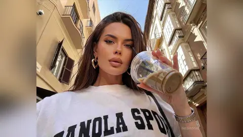 InesMoore poses outdoors in a white t-shirt, holding a cold coffee drink while looking at the camera with long brown hair and heart-shaped earrings.