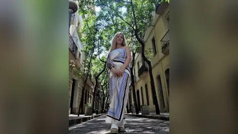Blonde woman in a striped maxi dress standing on a sunny urban street lined with trees and buildings.