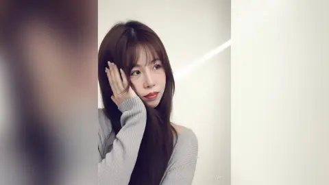Young woman with long brown hair wearing a grey ribbed sweater in a bright, minimalist setting.