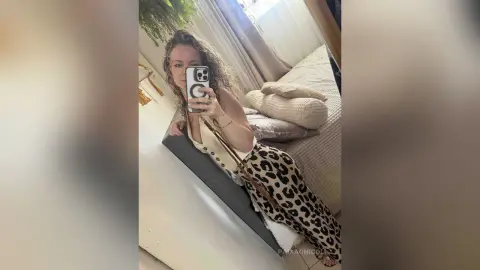 Smiling woman in a white tank top and leopard print pants taking a selfie in a bright bedroom.