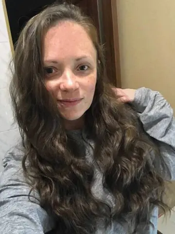 Smiling woman with long wavy brown hair wearing a cozy grey sweatshirt in a casual indoor setting.