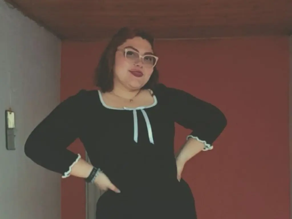 Smiling woman in a black long-sleeve dress with white trim posing against a red wall in a cozy indoor setting.