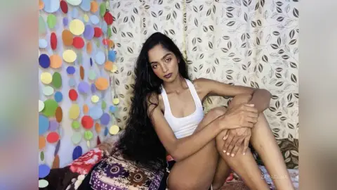 Young woman with long dark hair in a white tank top sitting against colorful paper decor and floral curtains.