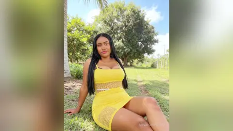 Smiling woman in a yellow summer dress sitting on green grass in a sunny tropical garden.