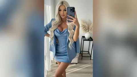 AlexaEvans poses in a blue satin dress and matching blazer, showing off her long blonde wavy hair and cleavage in a bright room.