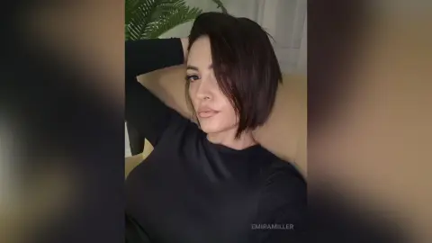 Young woman in a black long-sleeve shirt relaxing on a beige sofa with a calm, moody expression.