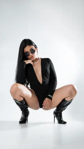 Stylish woman in a black bodysuit and sunglasses posing in high heel boots against a white studio background.