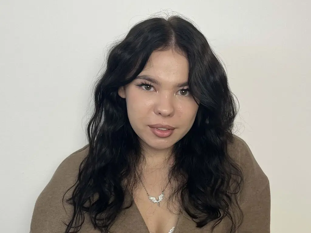 EmaRushlow looks at the camera with long wavy dark hair, wearing a brown top and a silver necklace against a white wall.