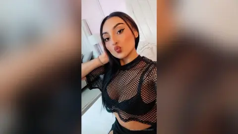 Young woman in a black mesh top posing with a playful expression in a bright bedroom setting.