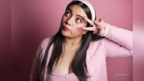Young woman in a pink fuzzy cardigan and headband posing playfully against a pink background.