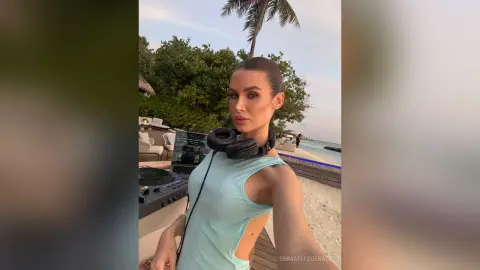 Young woman in a light blue sleeveless top wearing headphones on a tropical beach deck at sunset.