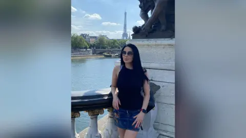 Stylish woman in a black tank top and denim skirt posing by the Seine River with the Eiffel Tower in Paris.