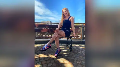 Blonde woman in a black mini dress and floral sneakers sitting on a bench under a blue sky.