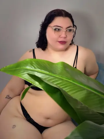 Curvy woman in black lingerie posing behind a large green leaf in a soft, moody indoor setting.