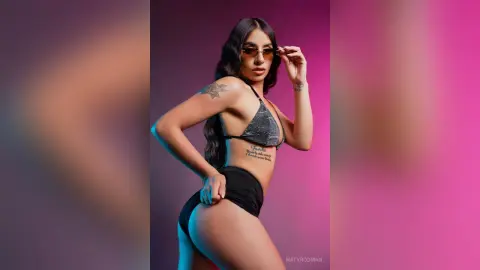 Confident woman in a black bikini and sunglasses posing against a purple studio background.